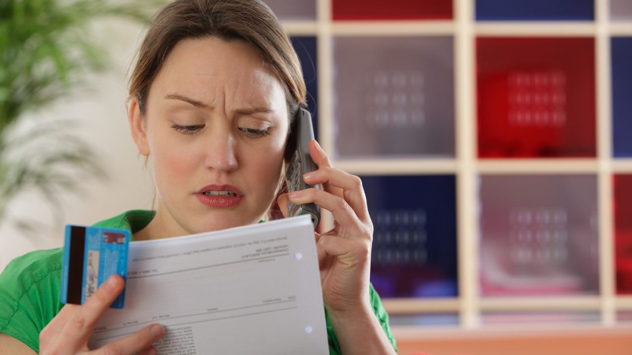 Disgruntled young woman holding a credit card and a bill and talking on the phone 