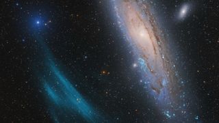 A bright galaxy hangs in space near a smear of blue nebulas gas and beside a mush smaller, more distant galaxy.