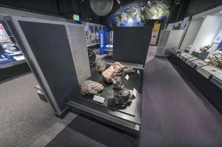 five large rocks sit on a display table in a museum, with photos of earth of and the moon on a wall in the background.
