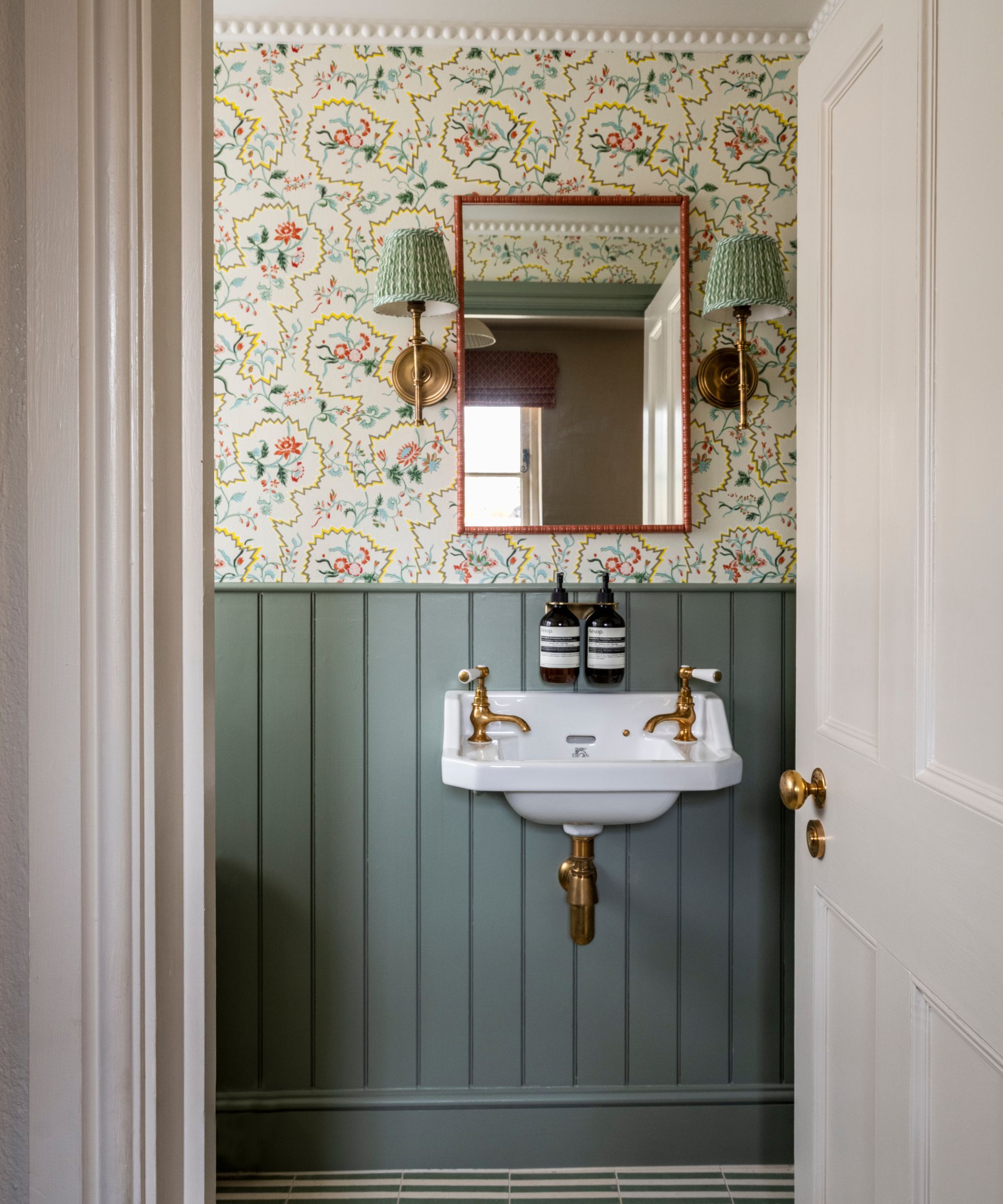 small powder room with striped green floor tiles, green panelling, a small vanity sink, floral wallpaper and a pink mirror flanked by gold and green sconces
