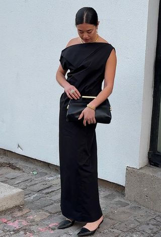 A woman standing outside wearing a black off-shoulder gown with gold jewelry, a black clutch bag, and black pointed pumps.