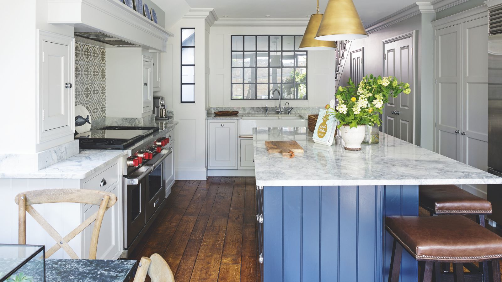 A blue and white kitchen with marble countertop, blue panelled island, brown wooden flooring and brown leather stools. Above the island are two gold pendant lamps. To the left is a silver stove with blue and white tiling above.