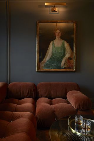 Image of a reddish-orange modular sofa in a slate-painted living room. There is a portrait painting of a woman hanging over the sofa and a gallery light shining onto the painting.