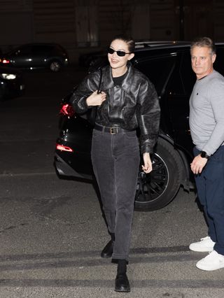 A photo of Gigi Hadid's off-duty model ensemble, which layers a black leather bomber with washed black jeans and lug sole loafers.