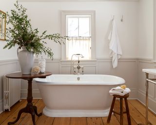 white bathroom with white roll top tub, wooden floor, wooden occasional table and light gray panelling