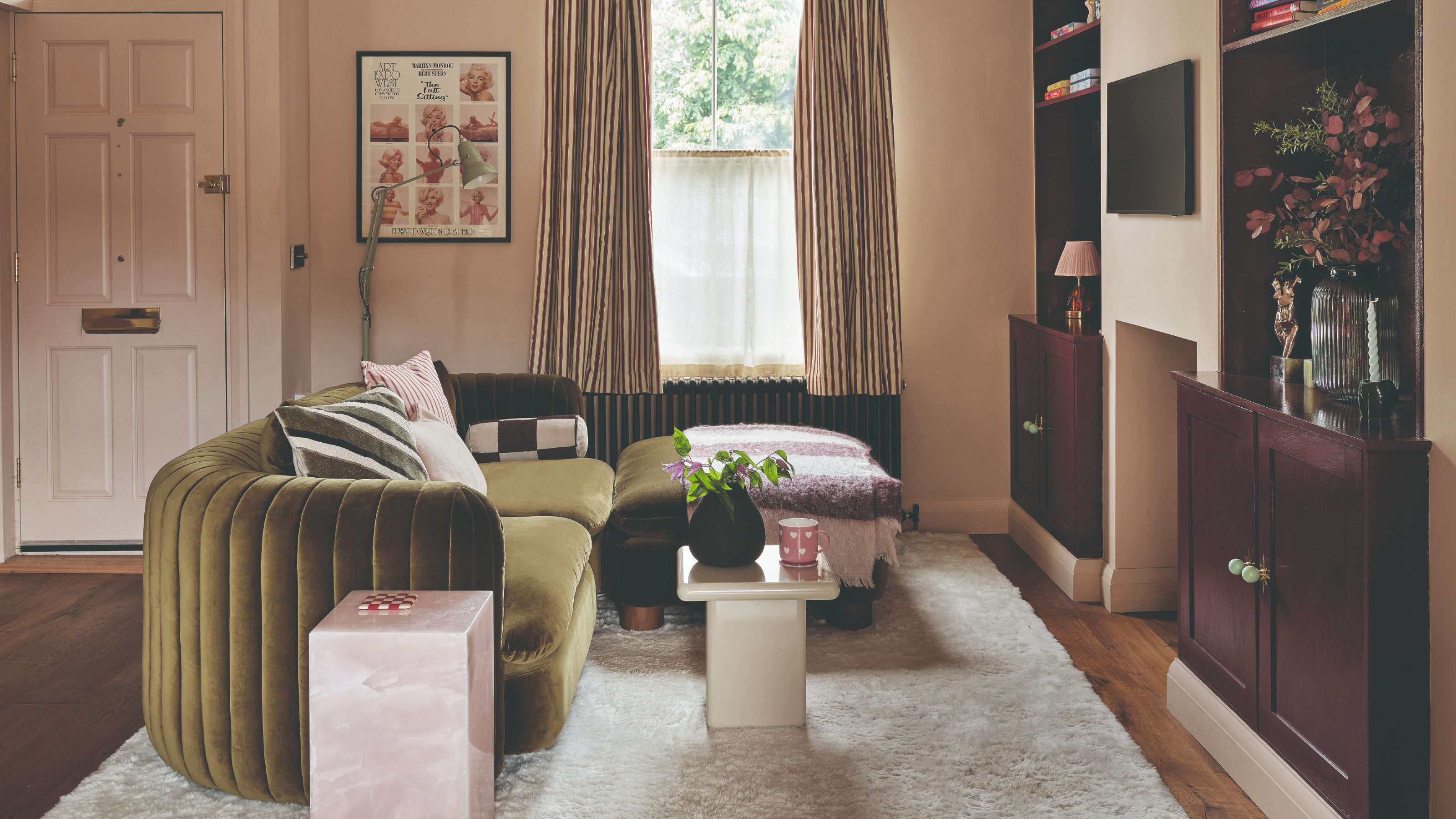 A small pink-painted living room with an olive green velvet curved sofa and burgundy bookcases built into alcoves