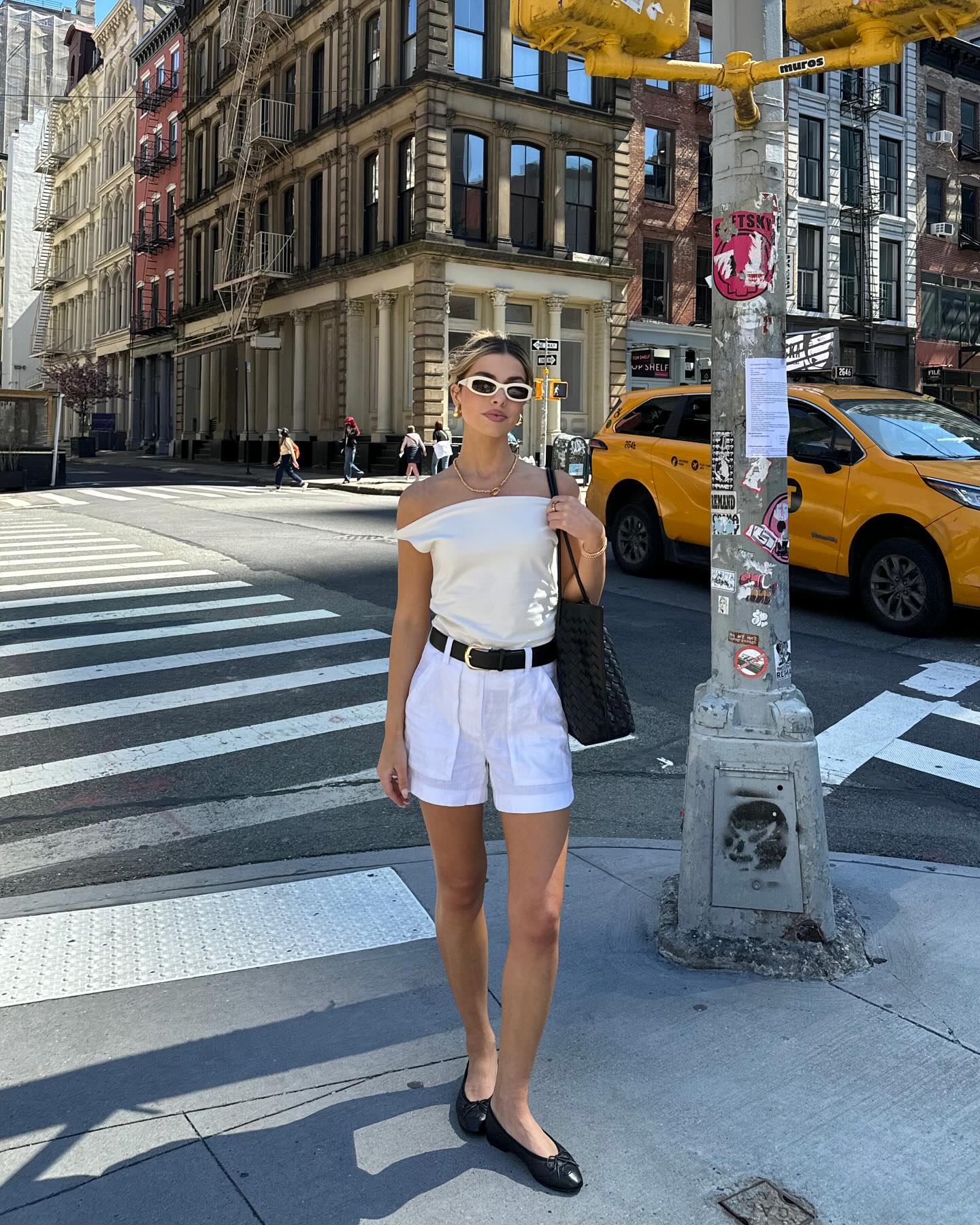 Woman wearing off-the-shoulder cream top, white shorts, and ballet flats shoe trend while posing on New York City intersection.