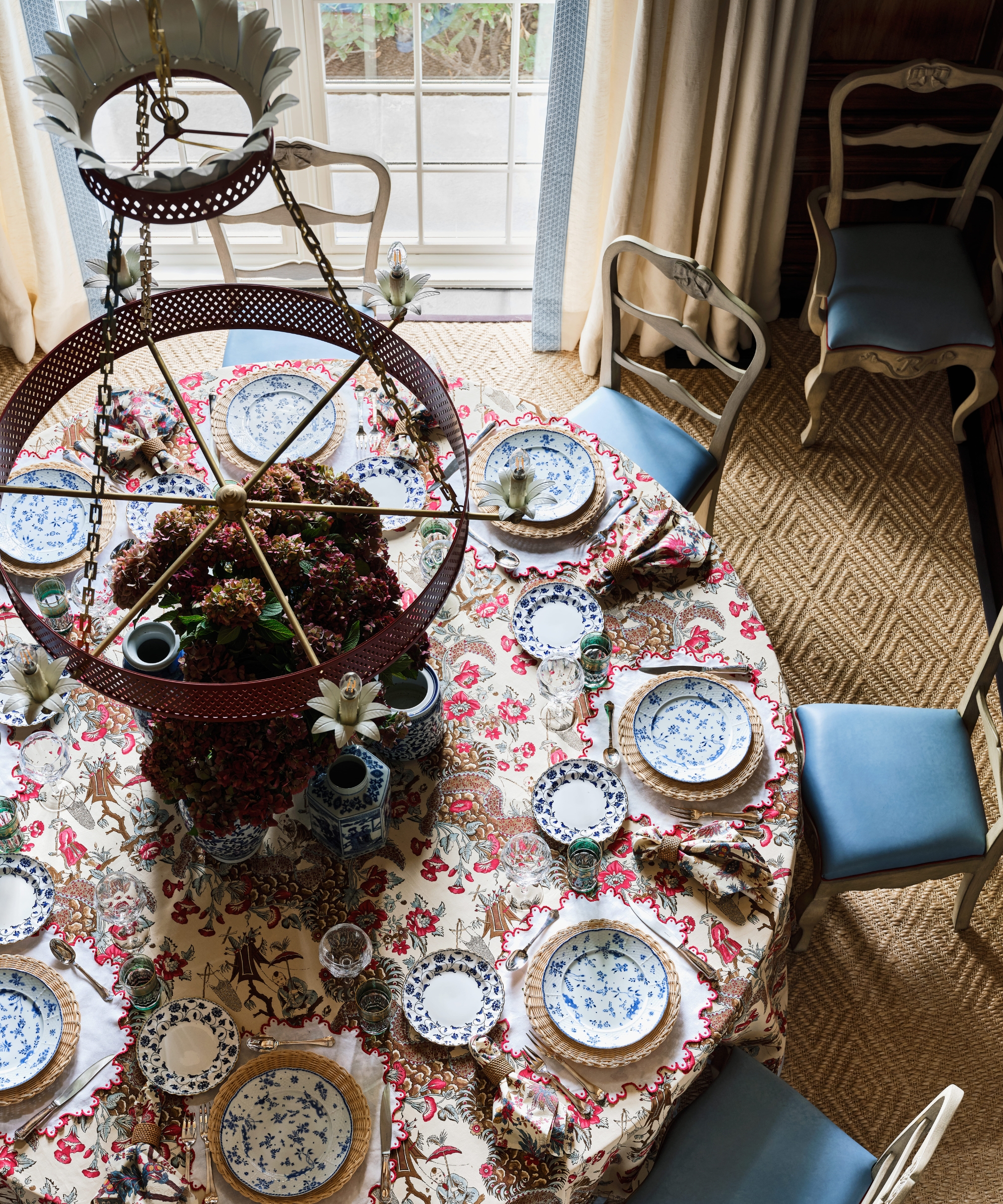 Overhead view of a round dining table layered with floral linen, blue-and-white china, hydrangeas and sky-blue leather chairs.