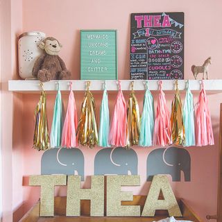 A pink-painted kids room with a display shelf and a glittered name sign spelling the name 'Thea'