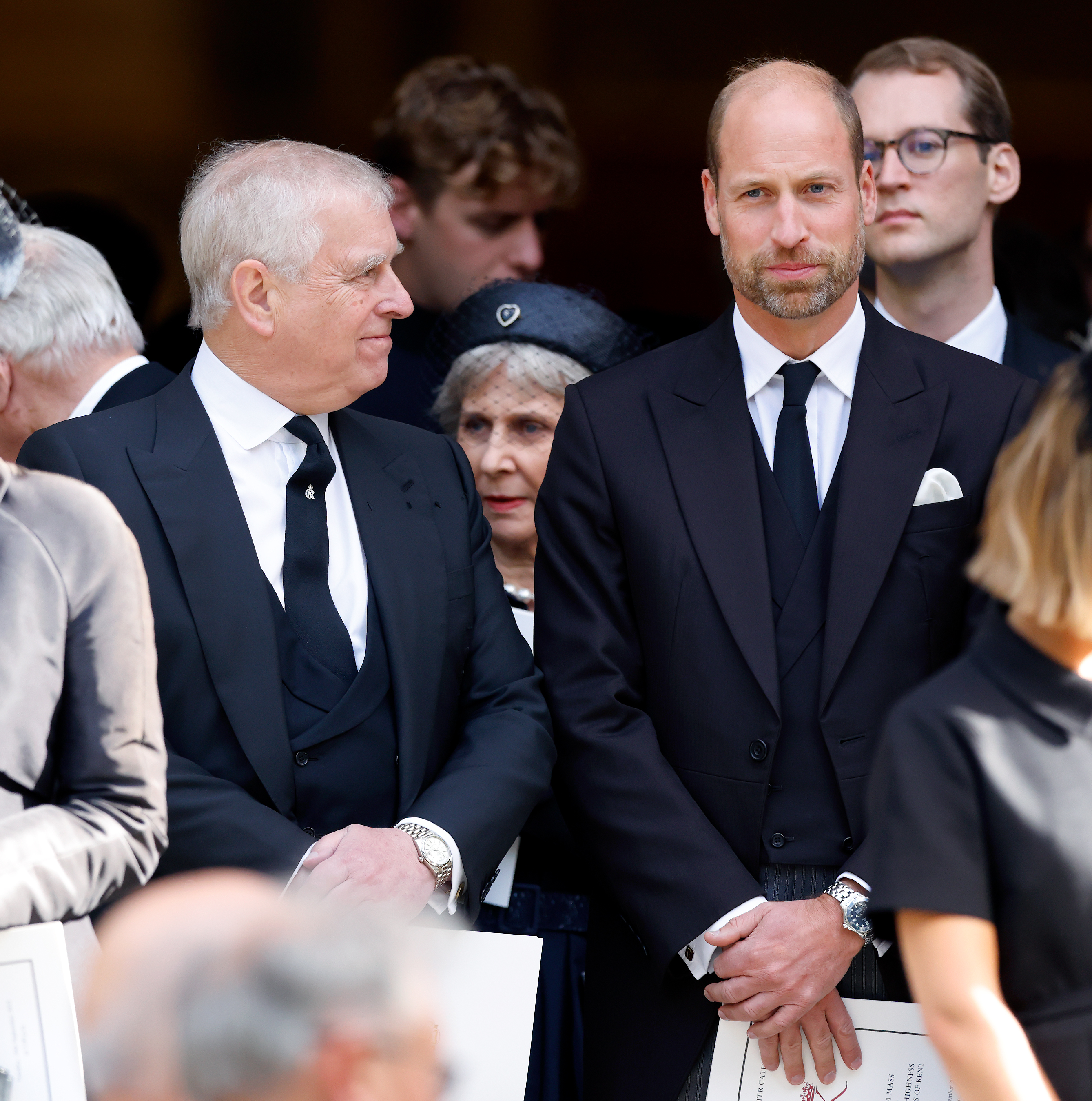 Prince Andrew smirking at Prince William, both are wearing dark suits standing in a group of people