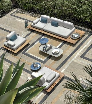 An outdoor setup with two single seaters and one 3 seater pebble-shaped light grey sofas, levitated on a large floating teak base with blue cushions. Blue round side tables and light grey rounded rectangle center table. All set in a courtyard with tiled flooring surrounded by green plants