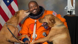 astronaut in a flight suit with two dogs jumping on him, on either side. behind is an american flag and the space shuttle