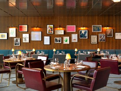 Interior view of the 9th floor restaurant at White City House. There is grey flooring, wood covered walls, colourful wall art with lights above, a row of blue chairs, round brown tables with lamps and tableware and deep red leather chairs