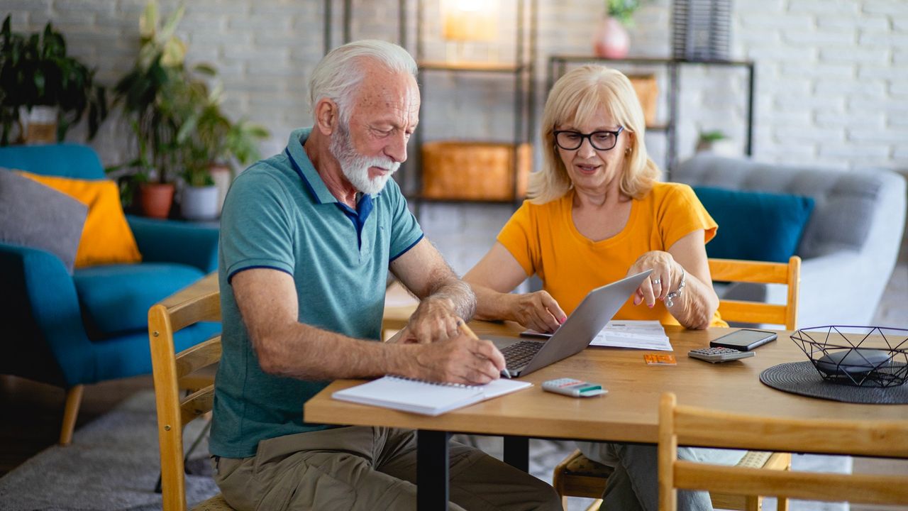 A retired couple sits at a table working on a computer.