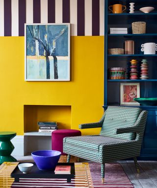 Colorful corner of a living room, yellow painted wall, fireplace, upholstered armchair, stiped painted design on upper wall, blue painted shelving unit, artwork on walls, books and ornaments on shelf