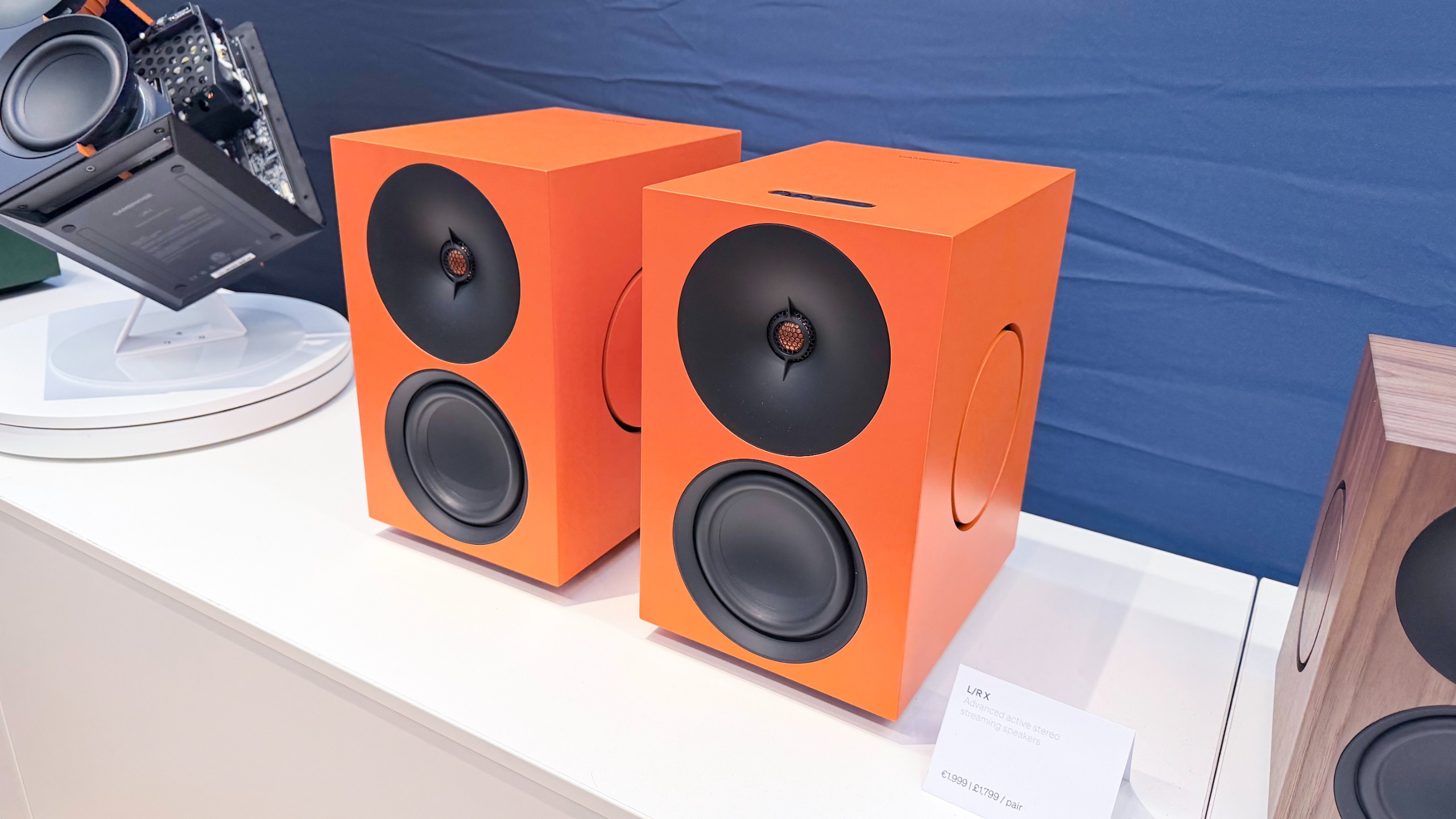 A pair of the Cambridge Audio L/R X speakers in orange, on a white surface against a blue fabric background.