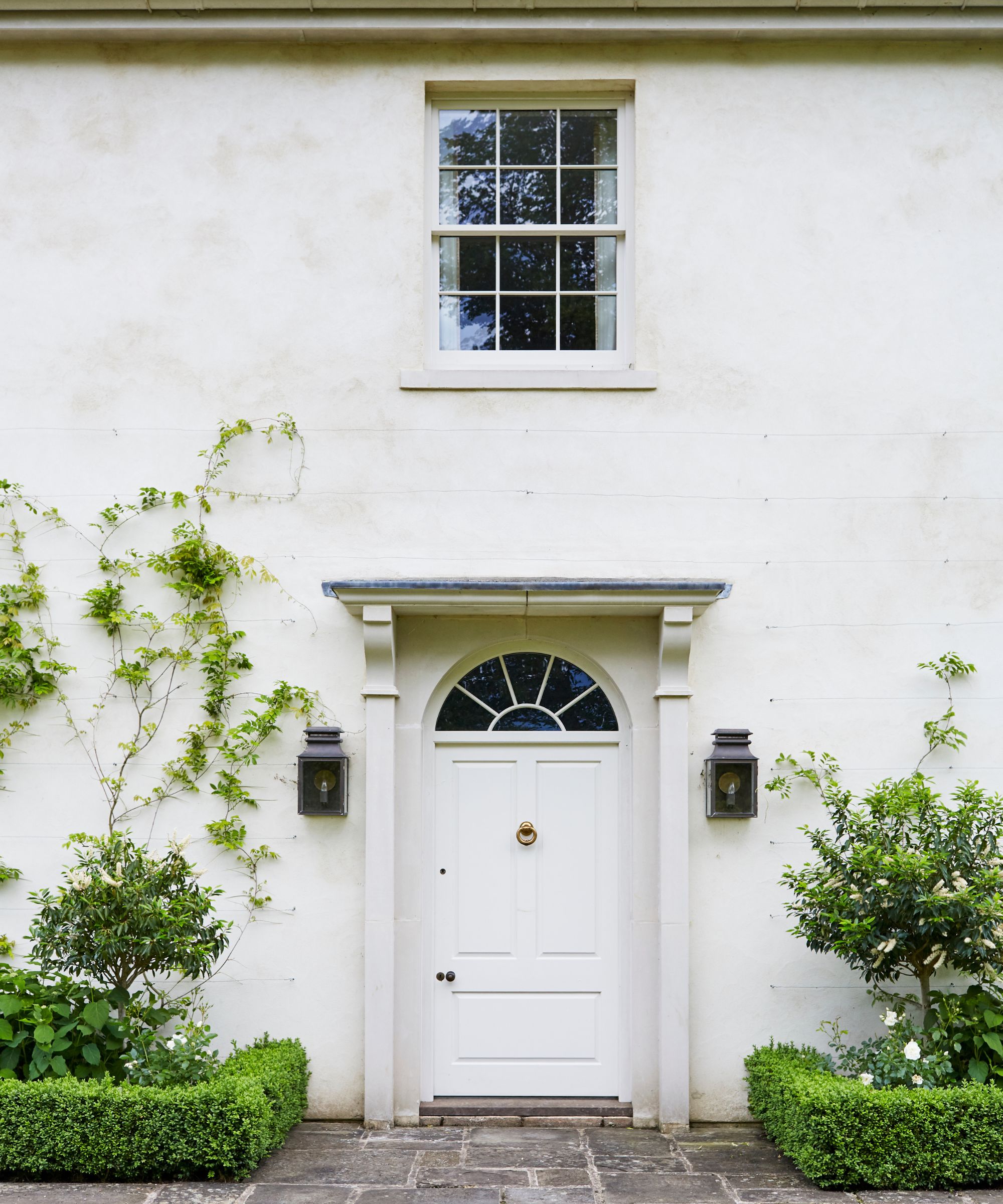 Georgian country house with white-washed exterior, grand front door with portico entrance, white front door, lantern sconces on either side and hedges with shrubbery