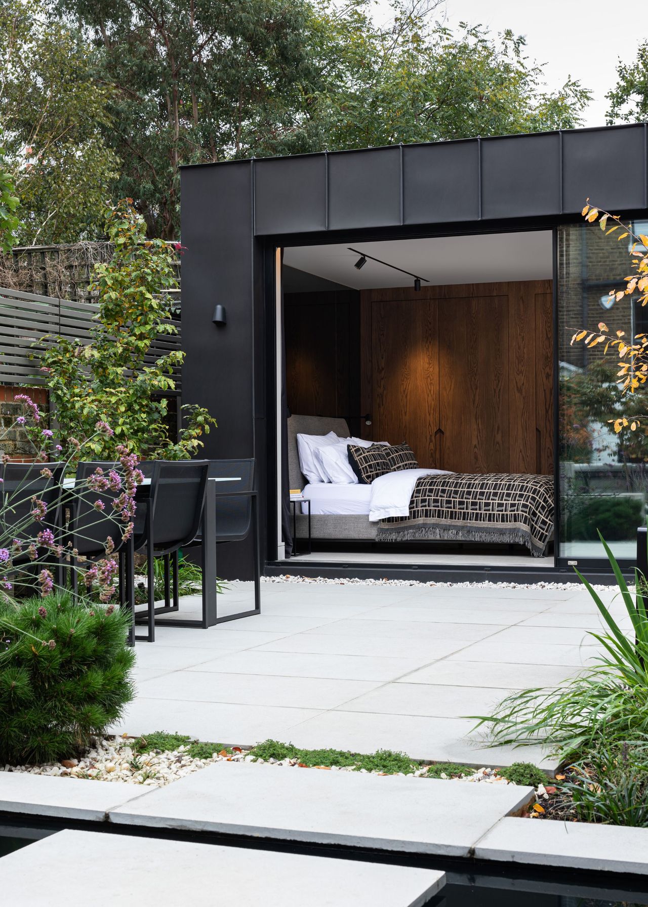 A patio with white stone flooring that also leads into a guest garden room where there is a view of the bedroom. In the patio area, there is also a dining table with chairs surrounded by lots of plants and flowers.