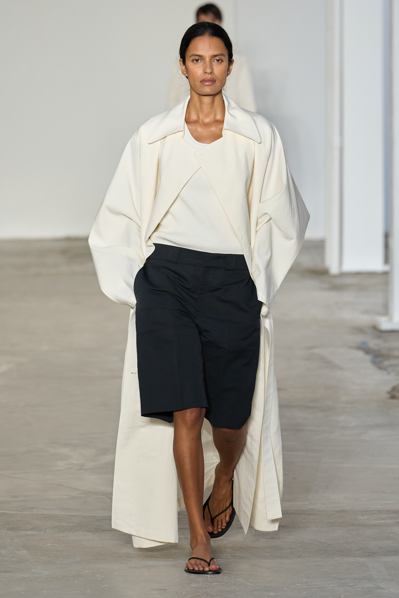 A FFORME model wearing a white coat, black shorts, and super thin flip flops.