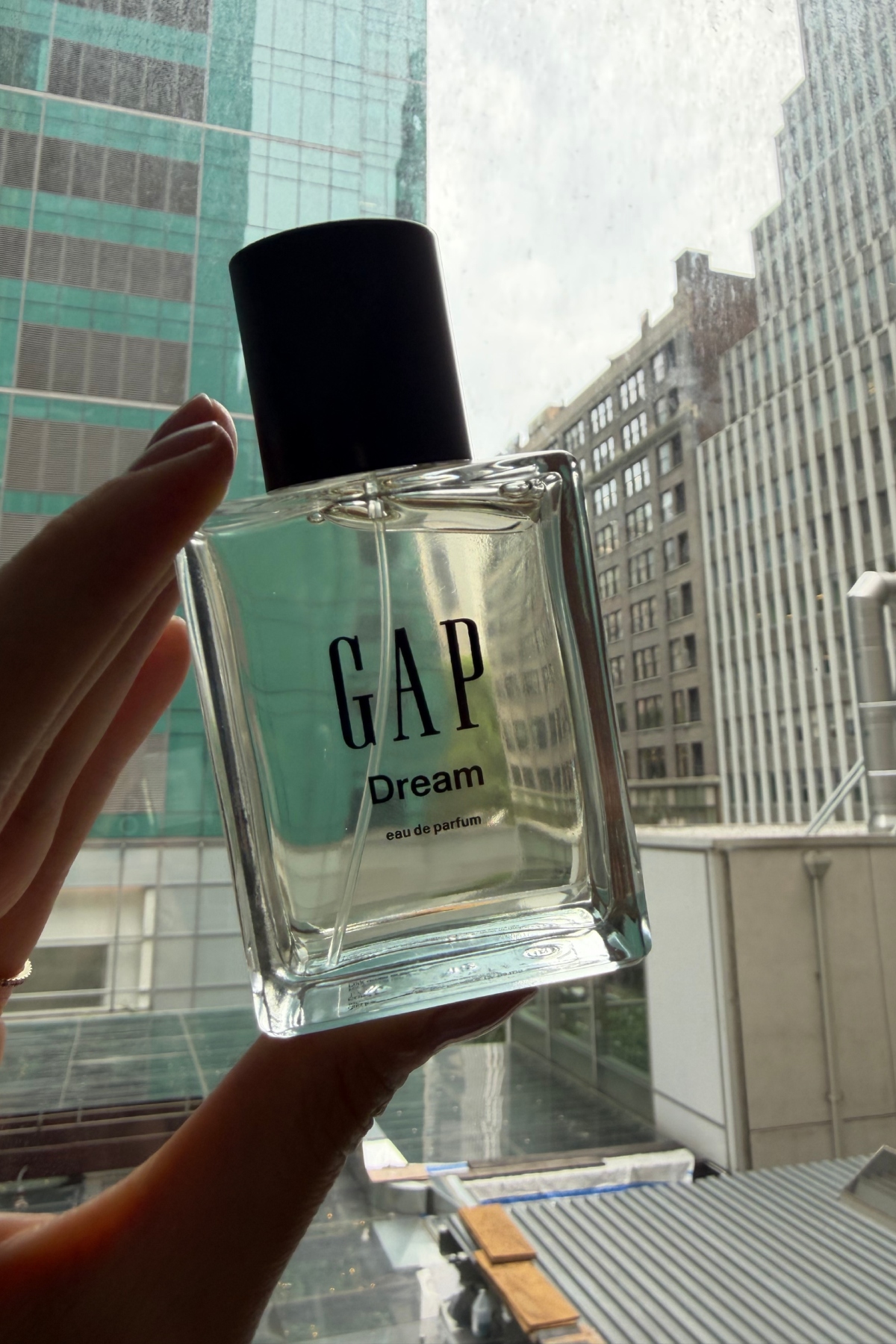 a hand holding a glass bottle of Gap Dream perfume up to the window