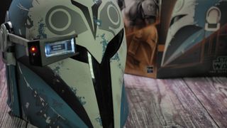 The Bo-Katan helmet with the rangefinder down sits on a wooden surface in front of the box