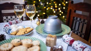 Festive spread, with bokeh lights in background. Mince pies, cracker and decorations in foreground with cookies and packaged Christmas pudding next to two wine glasses