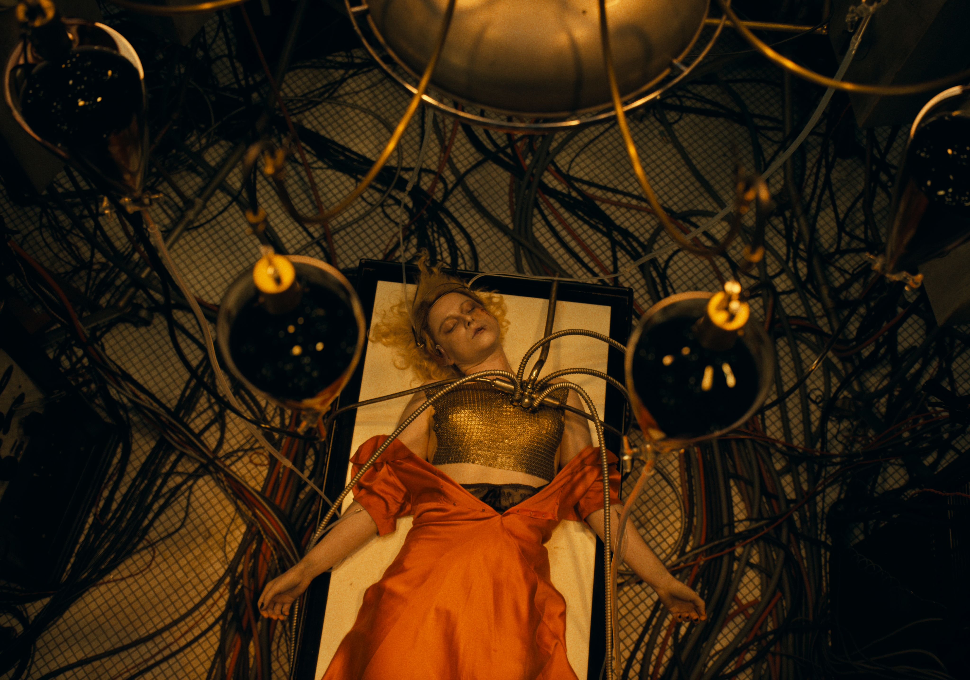 jessie buckley as the bride of frankenstein lying on an operation table with wires attached to her chest in a still from the movie the bride