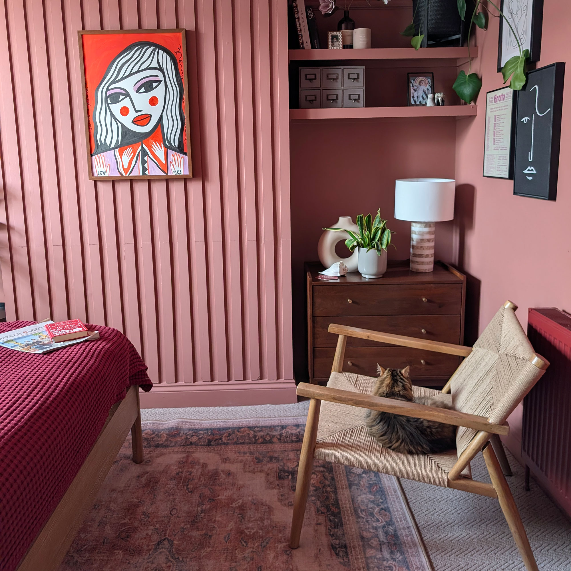 Pink panelled bedroom with graphic portrait of woman on wall, Persian style rug on floor, wood and woven armchair with cat sitting on it and dark brown chest of drawers in alcove