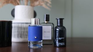 A selection of three men's fragrances on a wooden table. There is a green wall and dried flowers behind them.