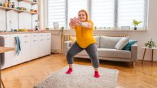 A woman performs a squat exercise in a home setting. Her knees are bent and her hands are held out in front of her. She wears a loose t-shirt, leggings and socks. Behind her is a sofa and windowsill with plants. Next to her is a kitchen unit with a sink and shelving.