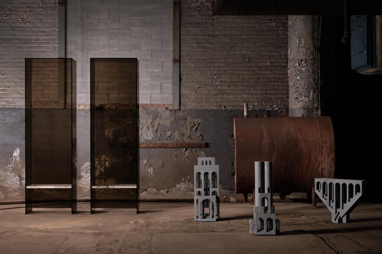 To the left, we see two chairs, made of see-through material in brown, with white marble-like seating. To the right, we see 3 objects, that look like models of different buildings or parts of them. It&#039;s all set in an industrial factory setting.