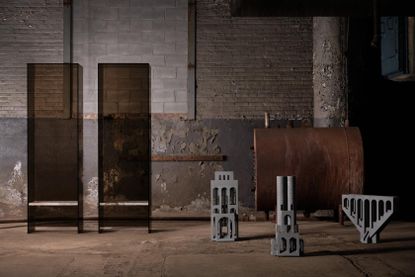 To the left, we see two chairs, made of see-through material in brown, with white marble-like seating. To the right, we see 3 objects, that look like models of different buildings or parts of them. It's all set in an industrial factory setting.