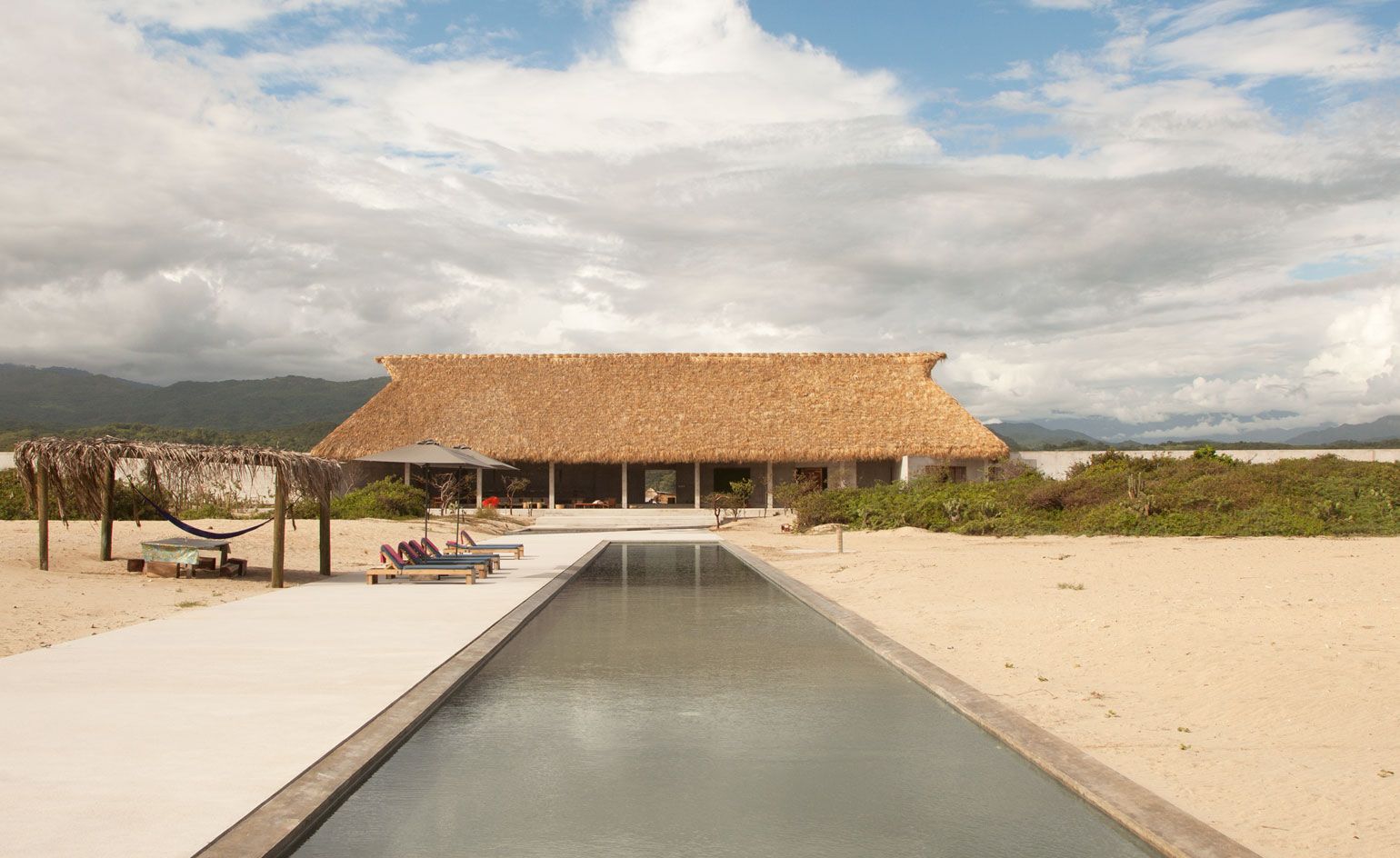Long swimming pool with concrete surround and loungers in front of a thatched palapa pavilion