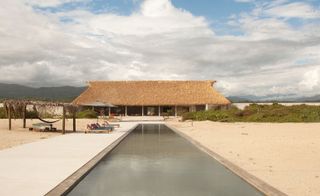 Long swimming pool with concrete surround and loungers in front of a thatched palapa pavilion