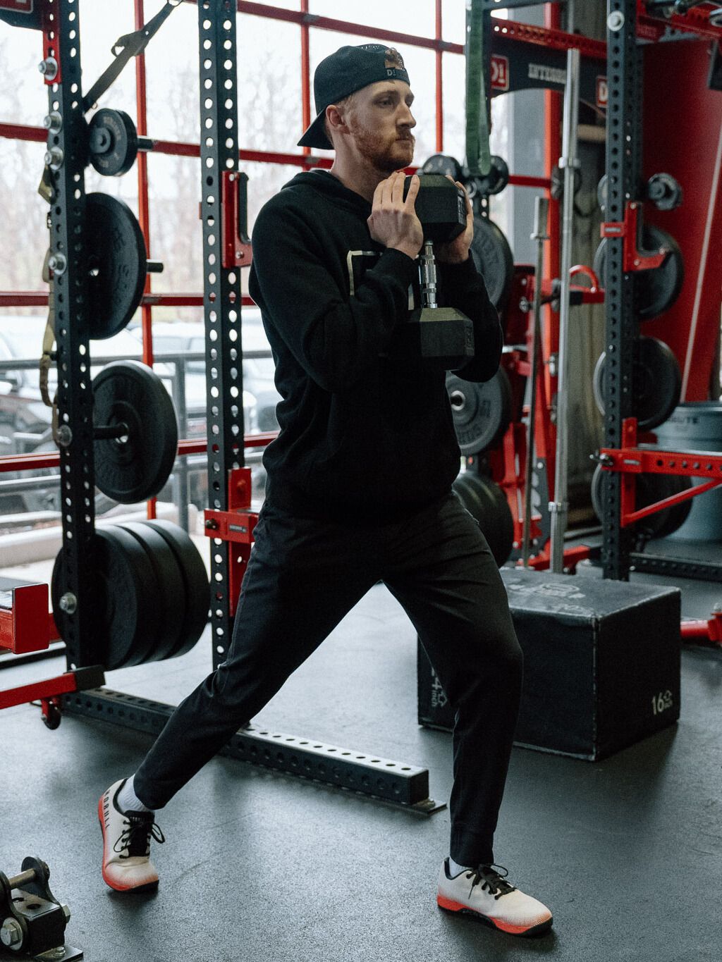 man in black hoodie and trousers performing a reverse lunge holding a dumbbell at his chest in a gym setting with a row of red squat racks behind him. 