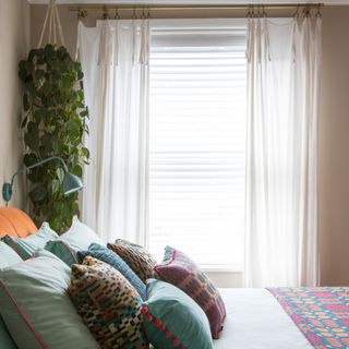 Beige painted bedroom with bed decorated with coloured cushions and hanging planter next to large window with blinds and sheer curtains