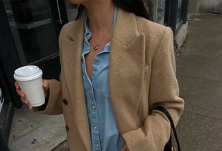 Sasha Mei wearing a camel coat, denim shirt, and jeans with a black tote bag.