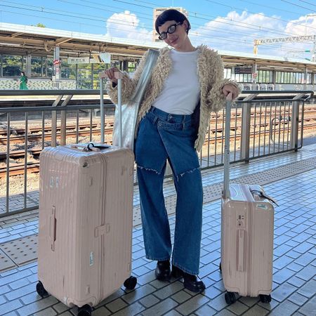 Alyssa wears a white tee, fur coat, jeans, black boots, and carries two pink suitcases.