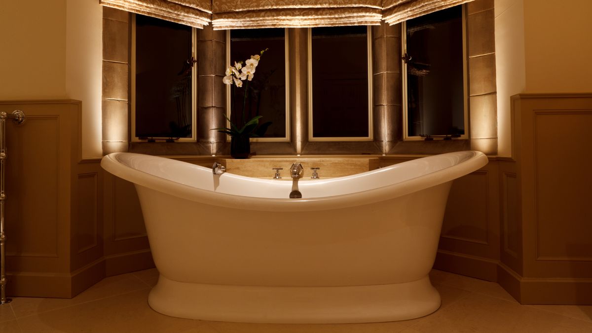 large white roll top bath in bay window with roman blinds, small lights recessed into windowsill and a light shining down into bath