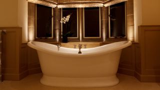 large white roll top bath in bay window with roman blinds, small lights recessed into windowsill and a light shining down into bath