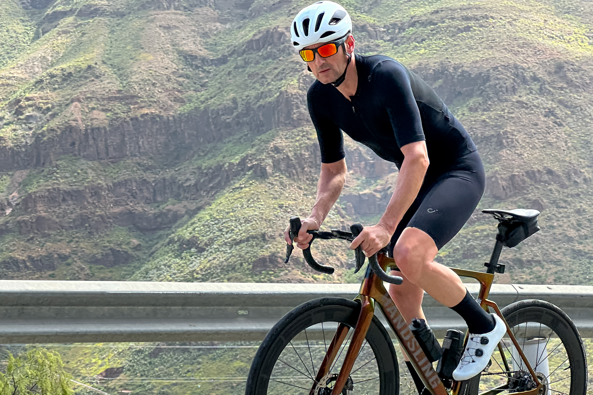man riding an orange bike up a hill out of the saddle wearing black shorts, white helmet, white shoes and black jersey