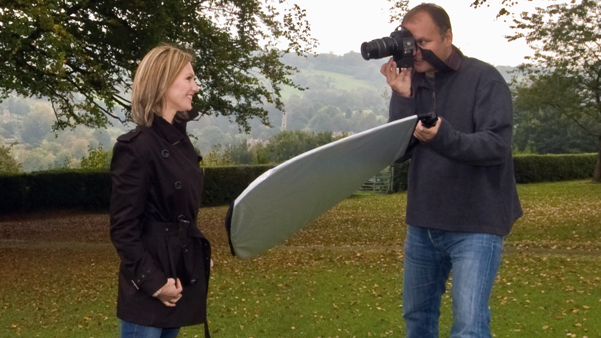 Photographer holding a Manfrotto TriGrip reflector in one hand and a DSLR in the other taking a portrait of a woman in a dark coat in a park in autumn