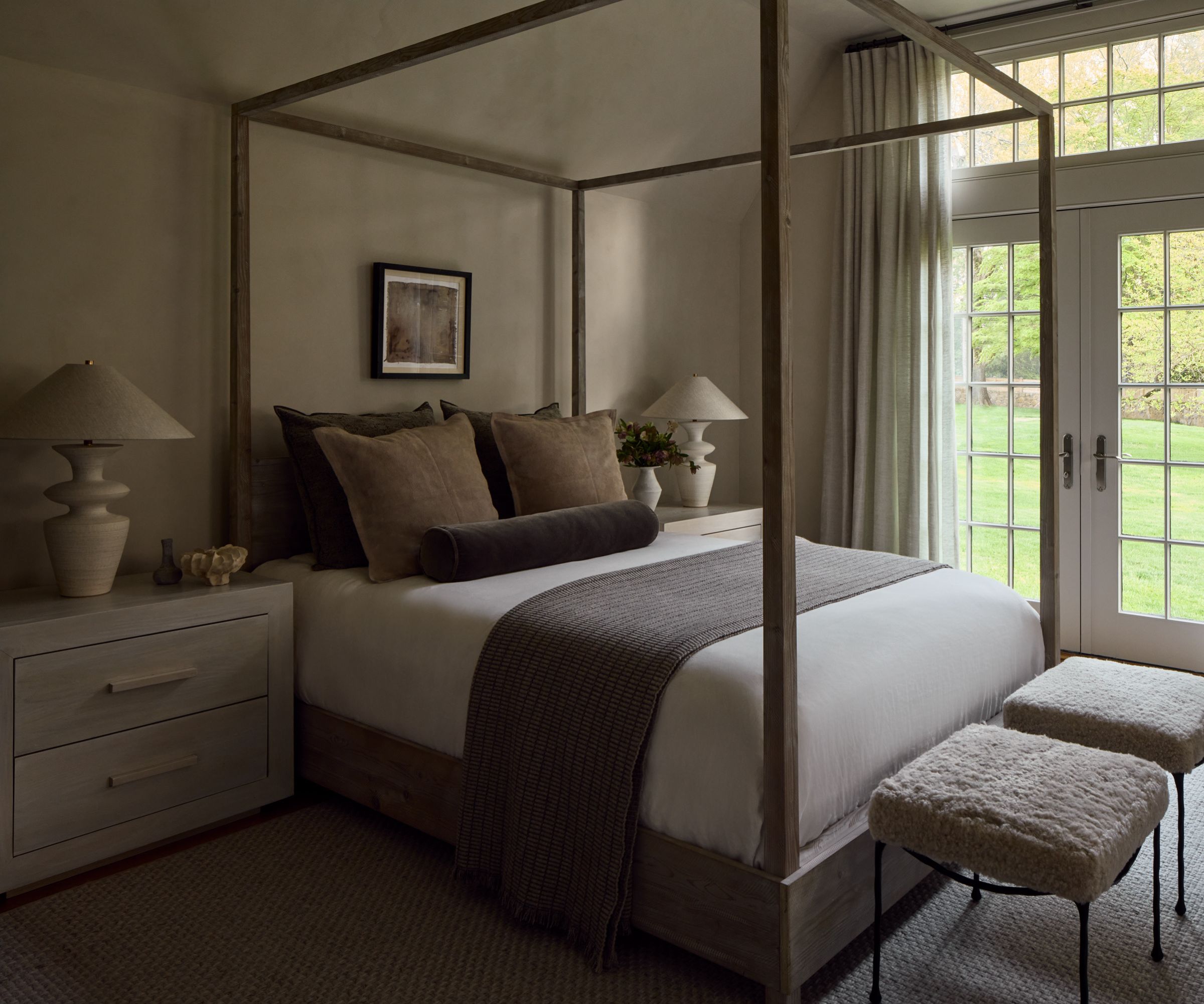A bedroom with taupe plaster walls and ceiling, a four-poster bed with neutral bedding, and French doors leading into a garden.