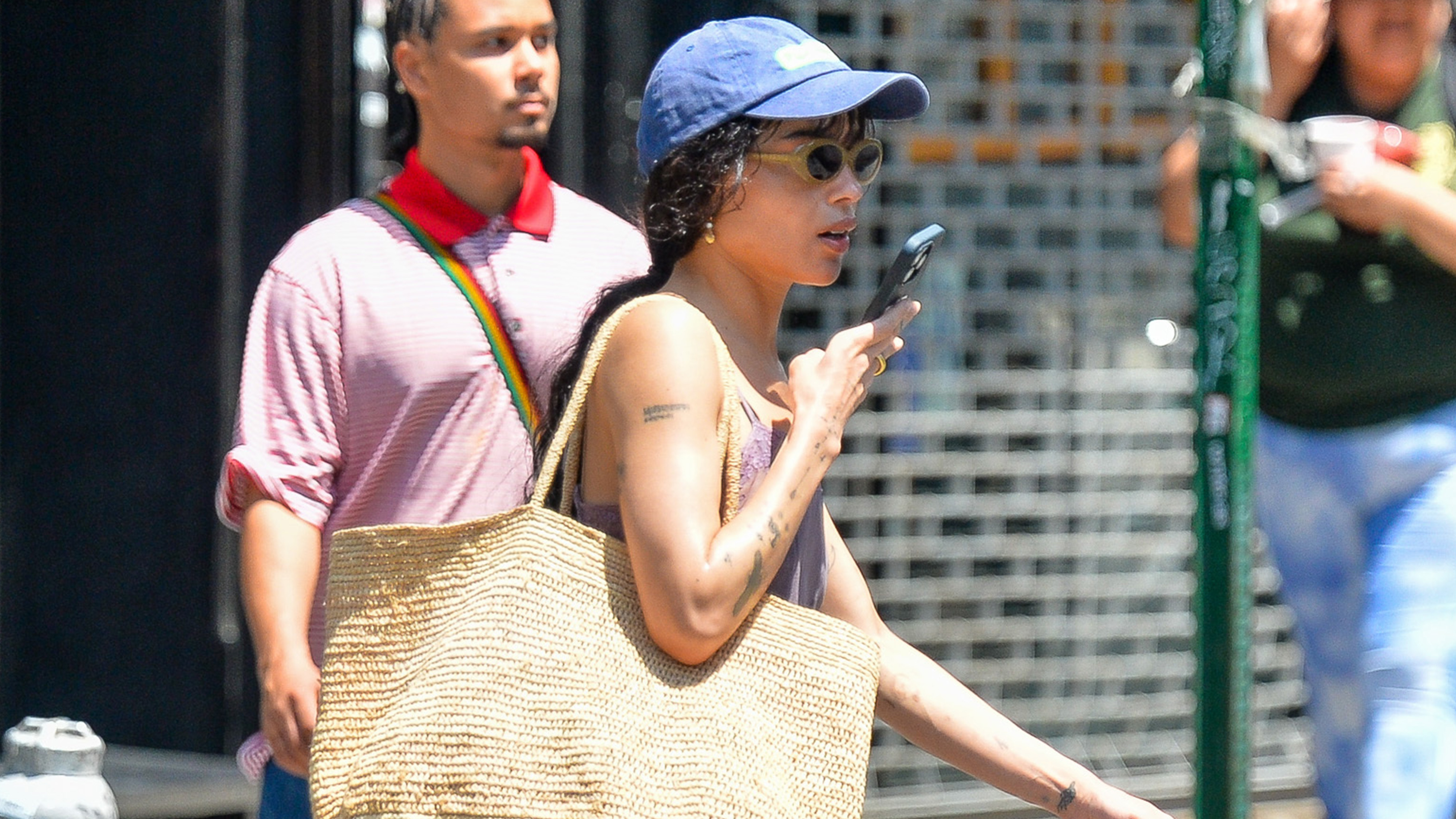 Zoe Kravitz wears a blue baseball cap, lavender dress, suede sandals, and a raffia tote bag.