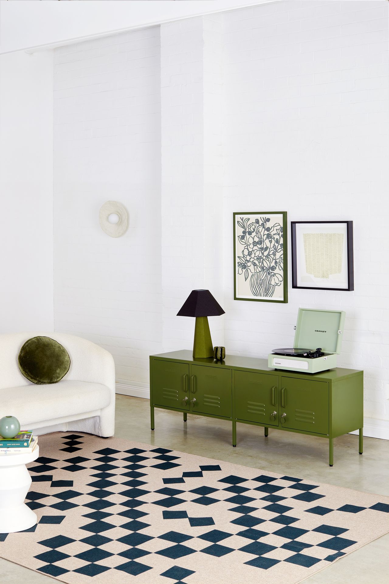 A white living room with a white sofa and a black and tan geometric rug. There is an olive green, low cabinet that resembles a locker that has a mint record player and an olive and navy blue lamp on it. There are two pictures hanging on the wall.