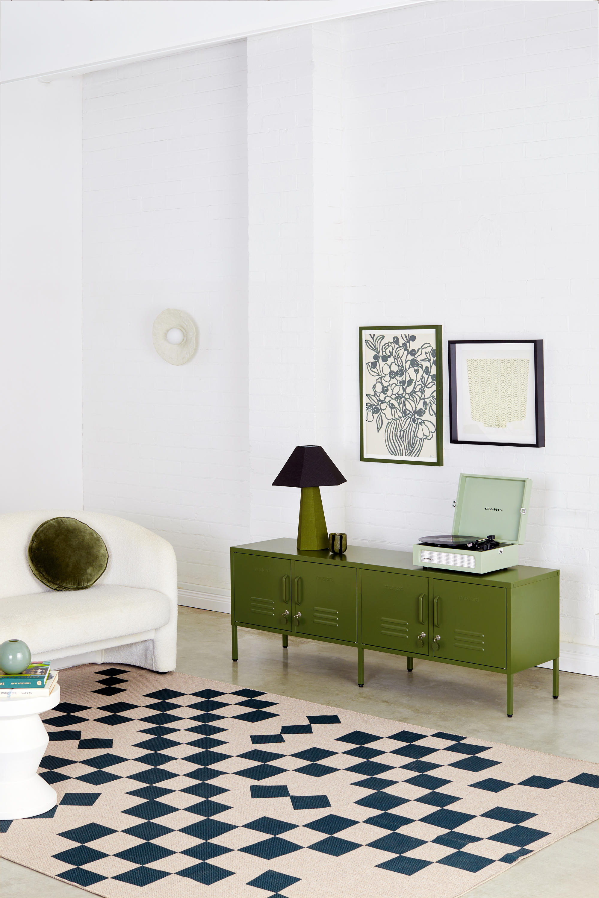 A white living room with a white sofa and a black and tan geometric rug. There is an olive green, low cabinet that resembles a locker that has a mint record player and an olive and navy blue lamp on it. There are two pictures hanging on the wall.