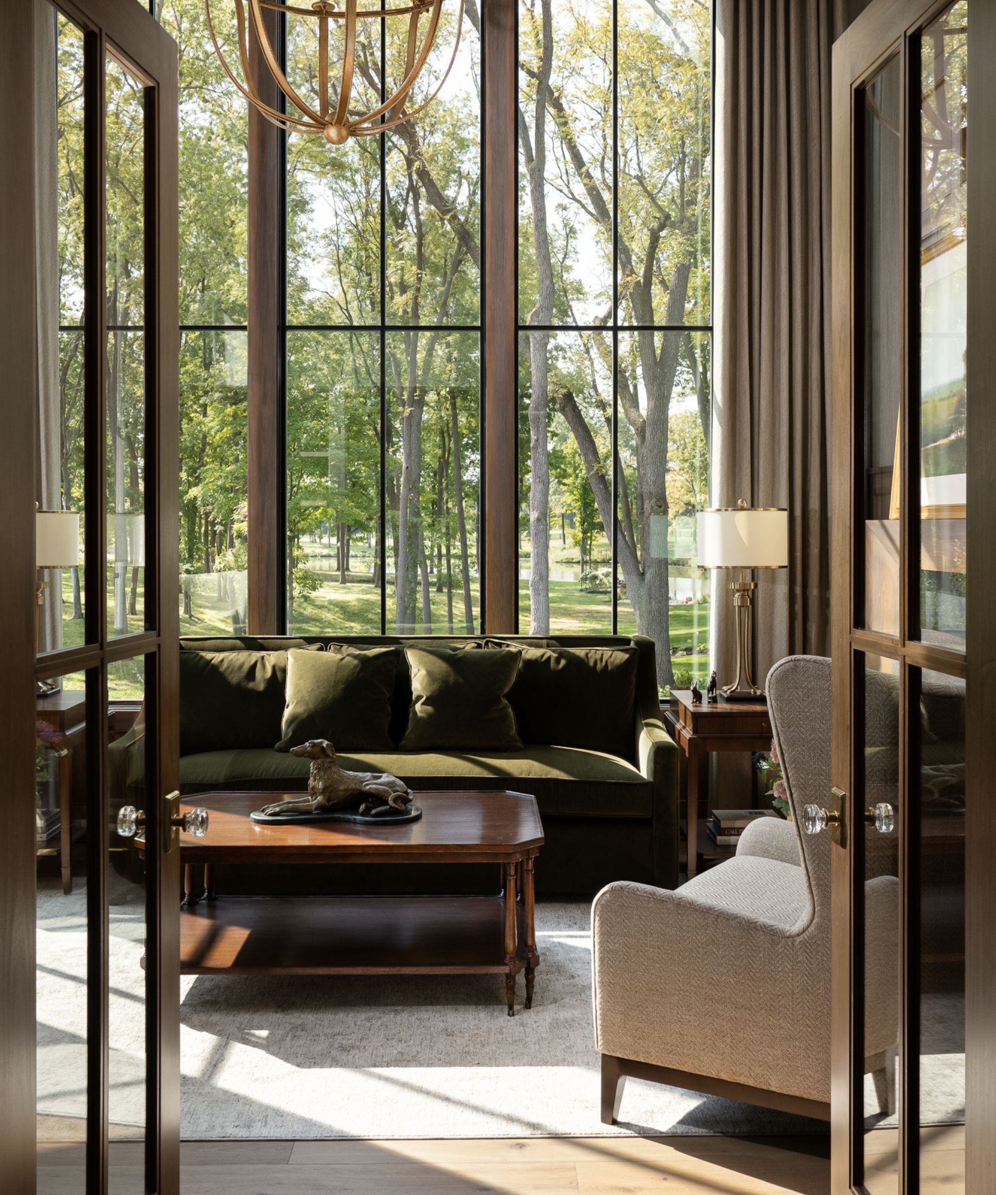 A high-ceilinged sitting room viewed through open French doors, featuring a moss-green velvet sofa, a wood coffee table with a dog sculpture, and massive windows overlooking a lush forest.