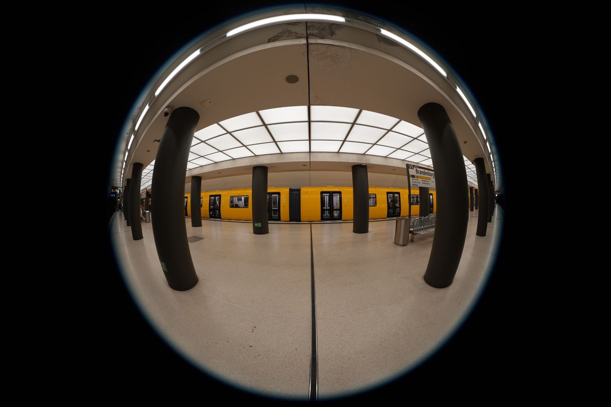 Circular fisheye view inside Brandenburger Tor U5 subway station in Berlin, with a yellow train behind black columns.