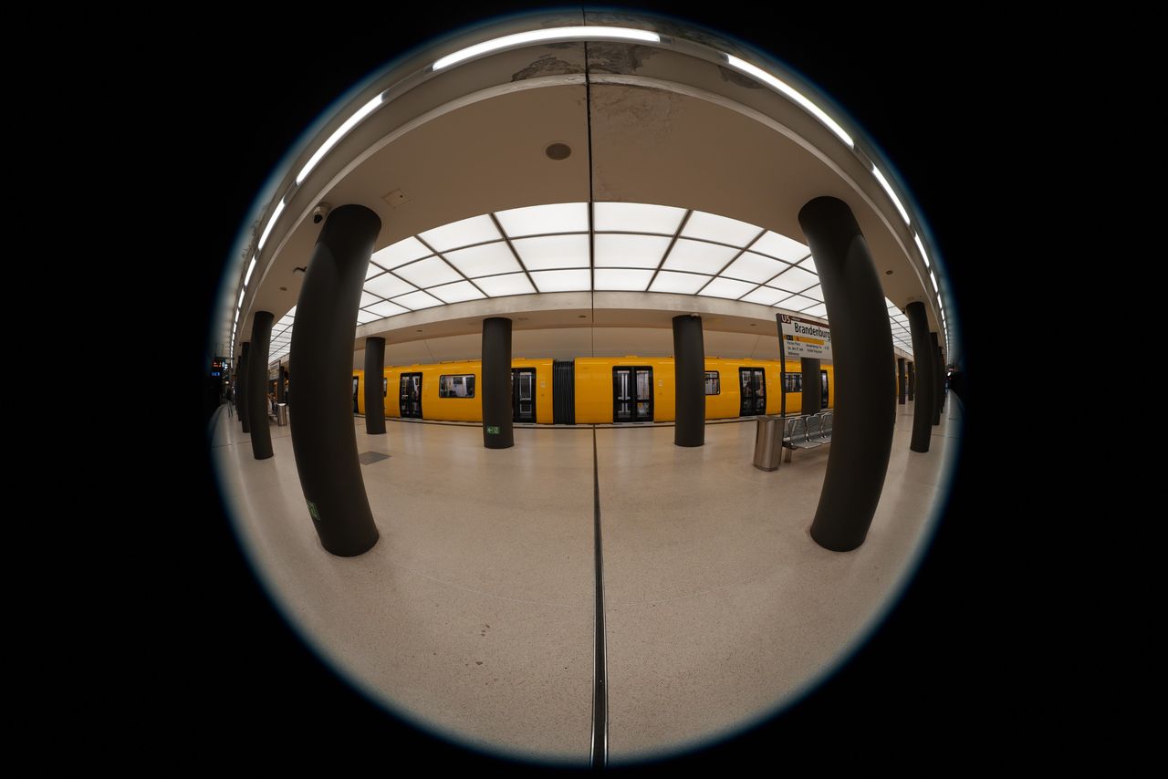 Circular fisheye view inside Brandenburger Tor U5 subway station in Berlin, with a yellow train behind black columns.