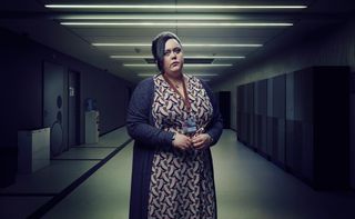 Sharon Rooney as Nina, standing in a school corridor wearing a pink and brown patterned dress and a long charcoal-coloured cardigan over it as well as a staff ID laminate.